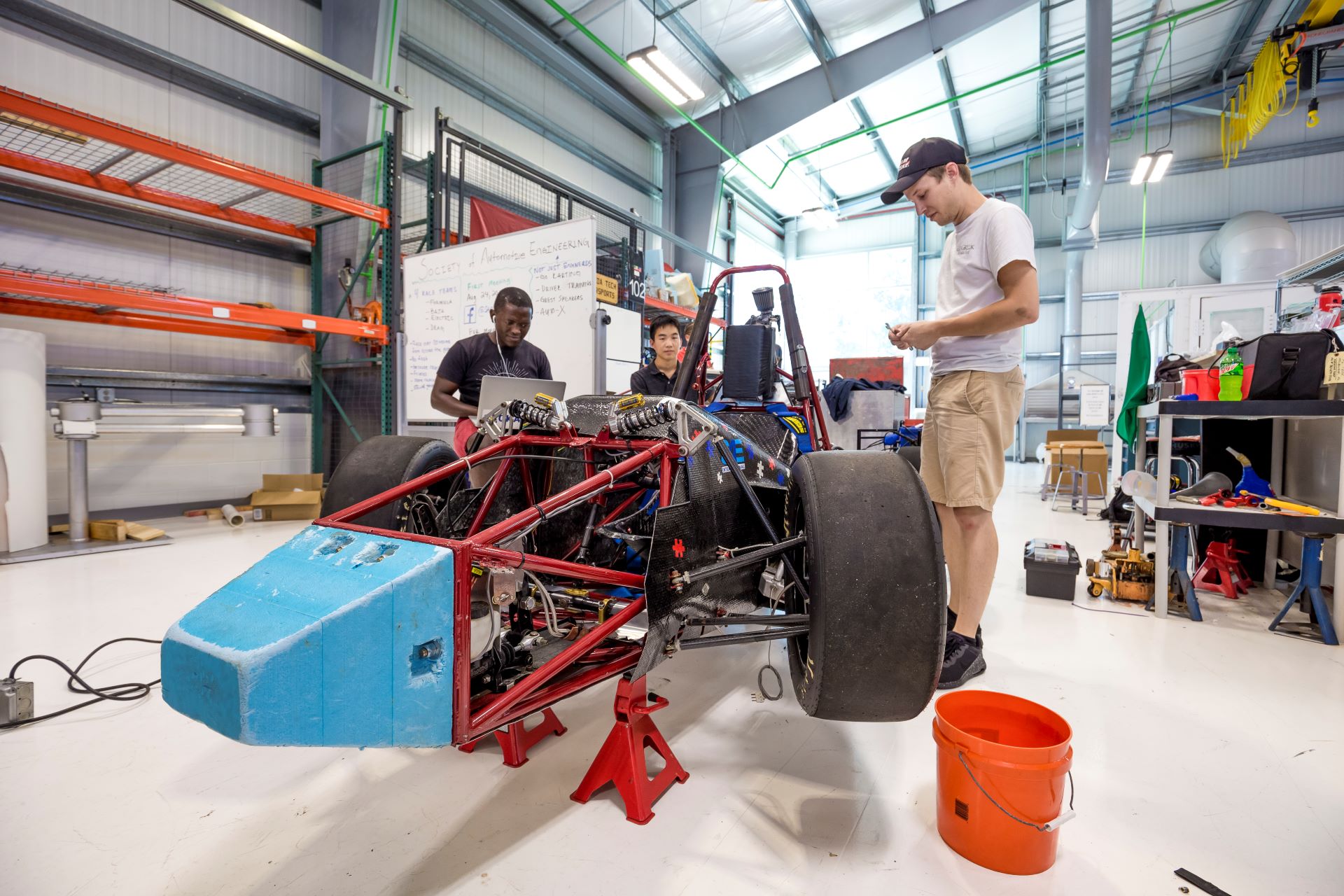 Three students working on a red formula racing car in a spacious engineering workshop, with tools and equipment scattered around